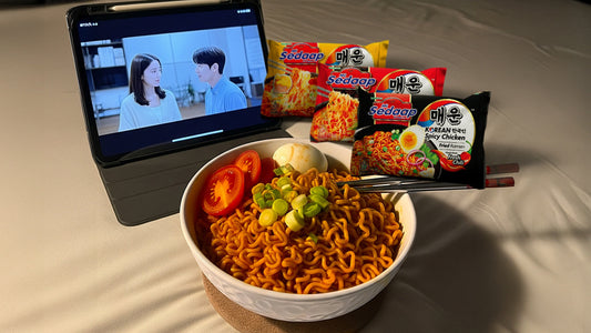 Mi Sedaap Korean Spicy Chicken ramen served with tomato, egg, and green onions, with a K-drama playing on a tablet.