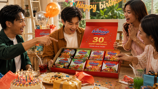 A group of friends excitedly celebrates a birthday around a table, cheering as a young man opens a red gift box filled with an assortment of Mi Sedaap instant noodle packs.