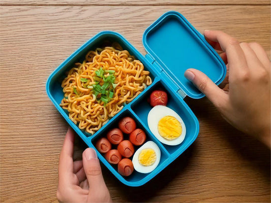 A blue bento box filled with noodles, mini sausages, and hard-boiled egg halves on a wooden table.