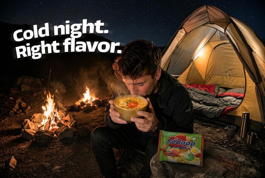 Man at a nighttime campsite holding a steaming bowl of Mi Sedaap Soto noodle soup, with a lit tent and campfire behind him.