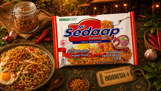 A plate of cooked Mi Sedaap fried noodles topped with a fried egg and chicken strips next to a packet of the noodles, surrounded by ingredients like chilies and garlic