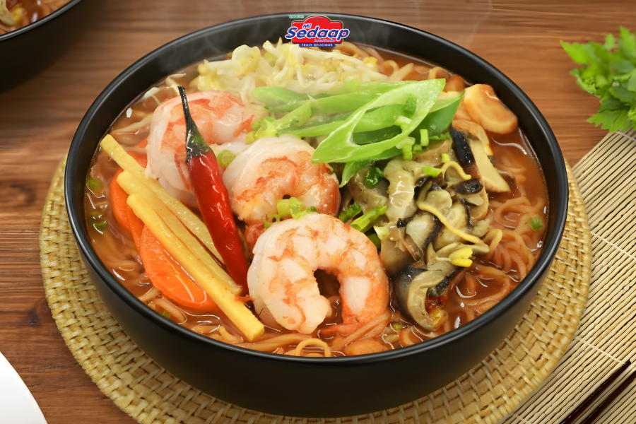 Bowl of Mi Sedaap shrimp noodle soup with spicy broth, shrimp, chili, cabbage, mushrooms, carrots, and green vegetables served on a woven placemat.