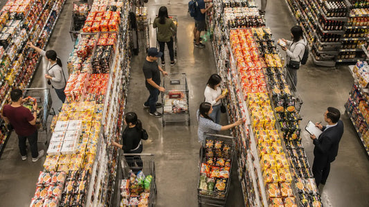 Aerial view of a grocery store instant noodle aisle with shoppers browsing colorful noodle packages and a buyer taking notes.